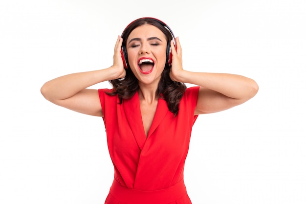 Glamorous girl in a red dress enjoys listening to music on headphones on white