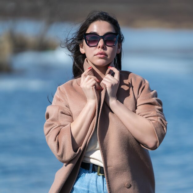 Glamorous hipster young woman in sunglasses dressed in beige coat standing outdoors against backgrou