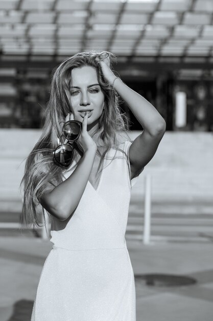 Glamorous long haired woman with perfect makeup holds sunglasses, posing at the sunny street. Monochrome color