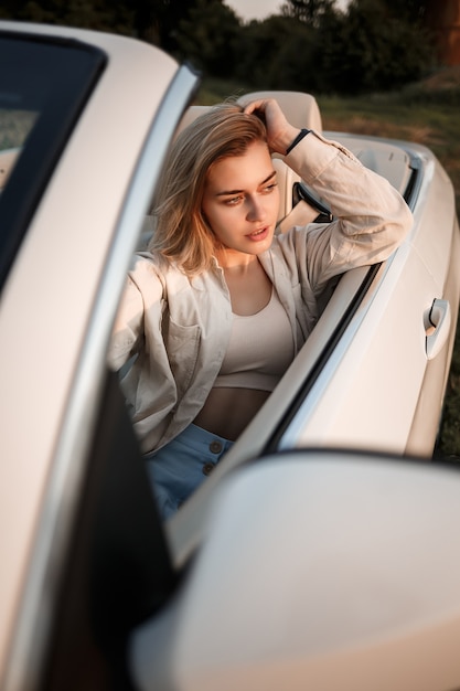 A glamorous luxury girl with blonde hair smiling while sitting on a white convertible. Young successful woman sitting in her white car