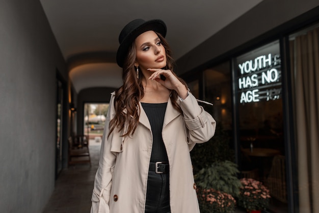 Glamorous stylish young pretty caucasian woman in fashionable autumn clothes with classic beige  coat and black hat walks on the street