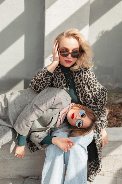 콘크리트 회색 벽 근처 거리에서 코트 청바지와 양복을 입은 세련된 선글라스를 착용한 매력적인 두 명의 아름다운 소녀는 친구와 함께 무릎을 꿇고 있습니다.