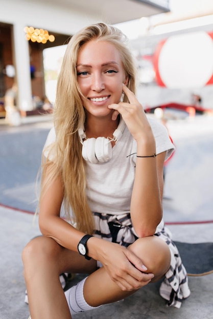 Photo glamorous woman in black wristwatch posing in skate park with smile adorable fairhaired skater girl in headphones laughing to camera