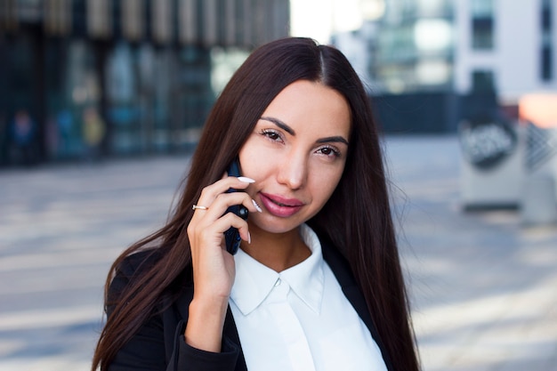 Glamorous young businesswoman  talking on her cell phone, looking at camera outdoor. Beautiful eastern caucasian brunette with big lips and long nails having conversation, call on smartphone.