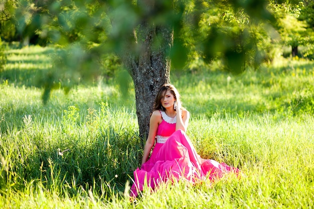 Glamorous young woman in a long pink dress sitting on grass under a tree