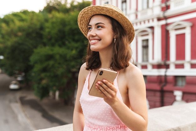 Affascinante giovane donna che indossa un cappello di paglia estivo che sorride e tiene in mano il cellulare mentre cammina per la strada della città