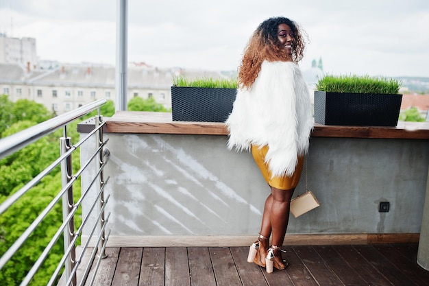 Glamour african american woman in yellow dress and white woolen cape with handbag posed at balcony