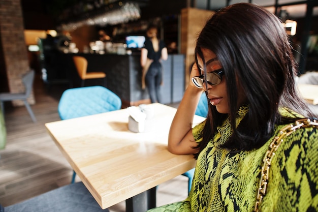 Glamour Afro-Amerikaanse vrouw draagt modieuze slangenjurk en zonnebril pose in restaurant.