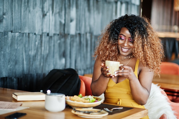 Glamour Afro-Amerikaanse vrouw in gele jurk zittend op restaurant met kopje thee.