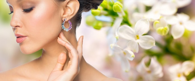 매력, 아름다움, 보석 및 고급 개념 - 천연 봄 라일락 꽃 배경 위에 귀걸이를 한 아름다운 여성의 얼굴 가까이