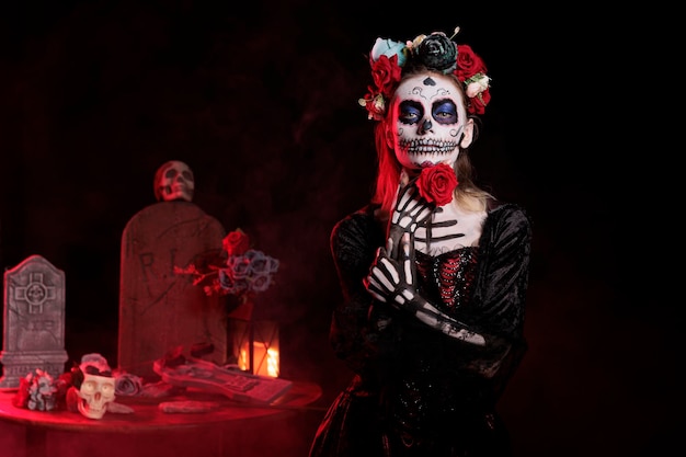 Glamour goddess of death posing with rose in studio, wearing flowers crown and santa muerte halloween costume. Looking like la cavalera catrina with body art and carnival look.