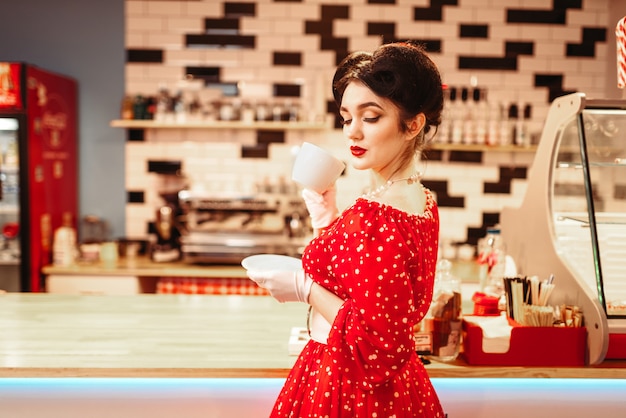 Glamour pin up girl with make-up drinks coffee in retro cafe, 50 american fashion. Red dress with polka dots, vintage style