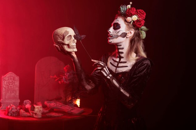 Glamour donna spettrale che si comporta in modo inquietante con il teschio in mano, con rose nere in mano e trucco spaventoso. assomiglia a santa muerte con corona di fiori e body art durante la santa celebrazione messicana.