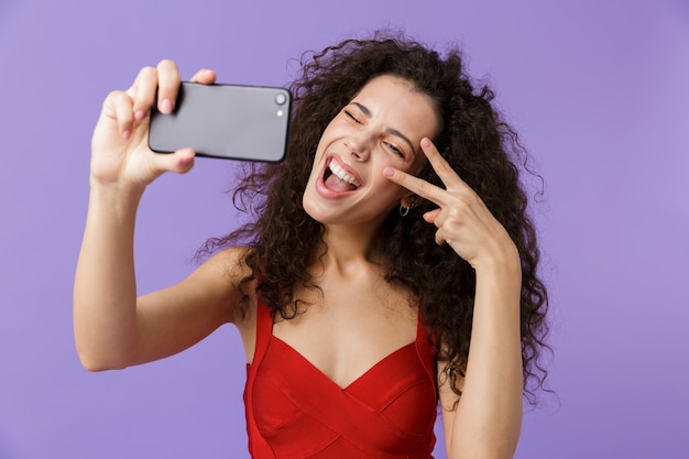 glamour vrouw draagt rode jurk selfie foto op zwarte mobiele telefoon, staande geïsoleerd over violette muur