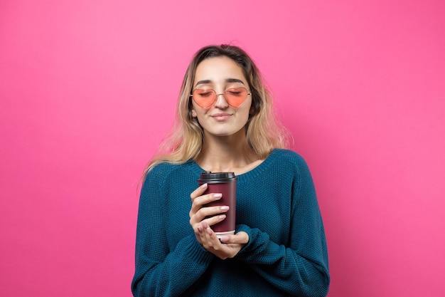Glamour vrouw in bril in een blauwe trui met een drankje van koffie op een roze achtergrond