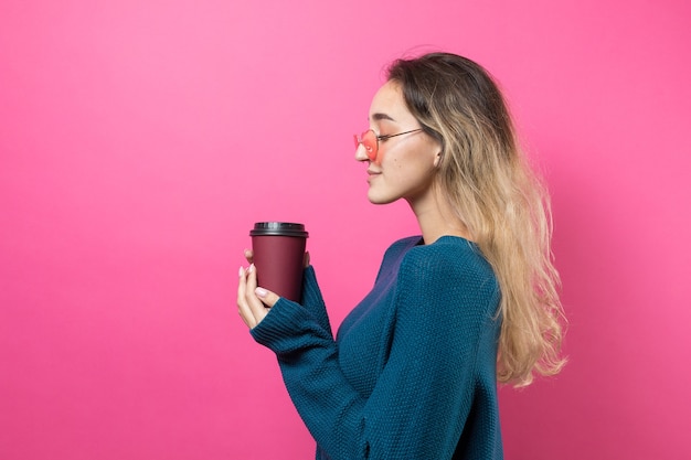 Glamour vrouw in bril in een blauwe trui met een drankje van koffie op een roze achtergrond