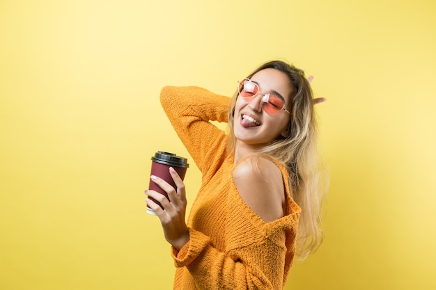 Glamour vrouw in glazen in een oranje trui met een drankje van koffie op een gele achtergrond