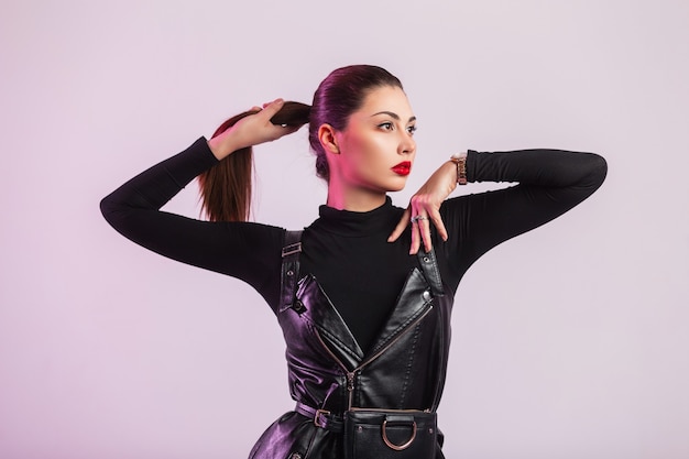 Glamoureuze moderne jonge vrouw mannequin met rode lippen met trendy kapsel in lederen modieuze zwarte jurk poseren in de buurt van vintage muur in de studio. Mooie brunette meisje in elegante kleding.