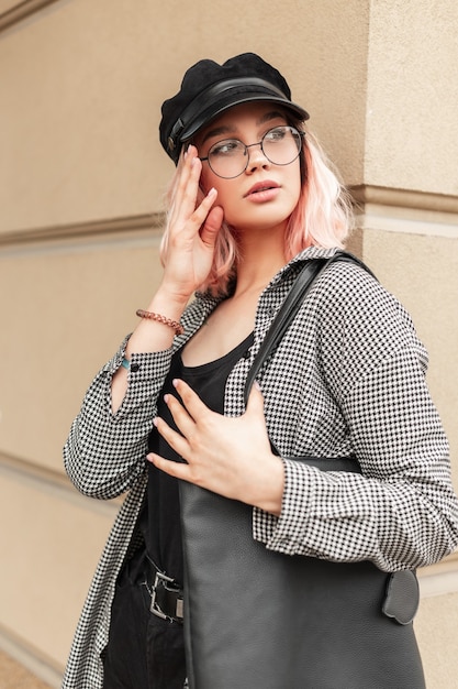 Glamoureuze mooie jonge vrouw met een bril en een hoed in trendy vrijetijdskleding met een tas die zich voordeed in de buurt van een beige muur in de stad