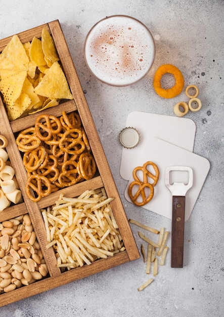 Glas ambachtelijk pils en opener met doos snacks op lichte keukentafel. Pretzel, zoute aardappelsticks, pinda's, uienringen met nacho's in vintage doos met openers en bierviltjes.