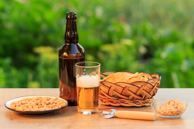 Glas bier en flesje bier op houten tafel met chips in rieten mand, pinda's in plaat en kom in natuurlijke groene onscherpe achtergrond