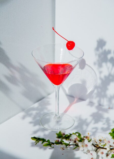 Glas cocktail en rode kers dichtbij tak van de boom van de bloesemkers in een witte hoek. Natuurlijk licht