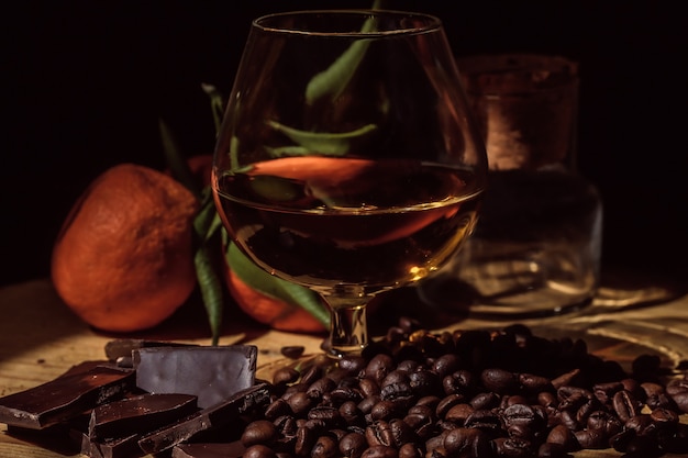 Glas cognac op houten tafel met nauwe chocolade, koffie en mandarijnen.