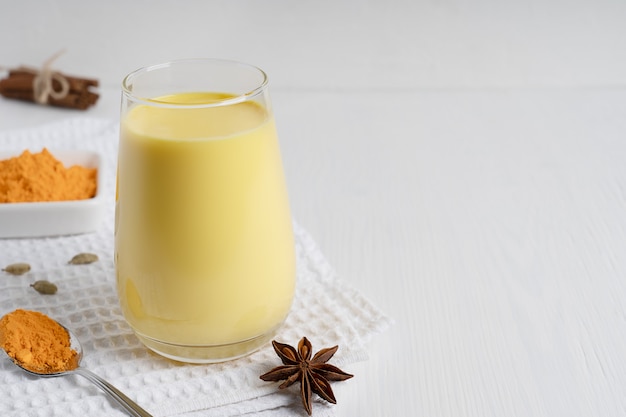 Glas gouden melk geserveerd met lepel kurkumapoeder en anijs op witte houten achtergrond