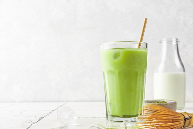 Glas ijskoude groene matcha latte met een rietje en een fles melk op een witte tegelachtergrond koude zomerdrank