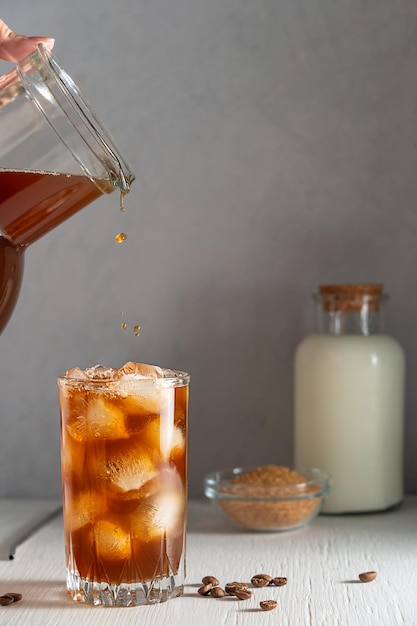 Glas koude ijskoffie met ijsblokjes en drinken uit een pot geserveerd op een witte houten tafel