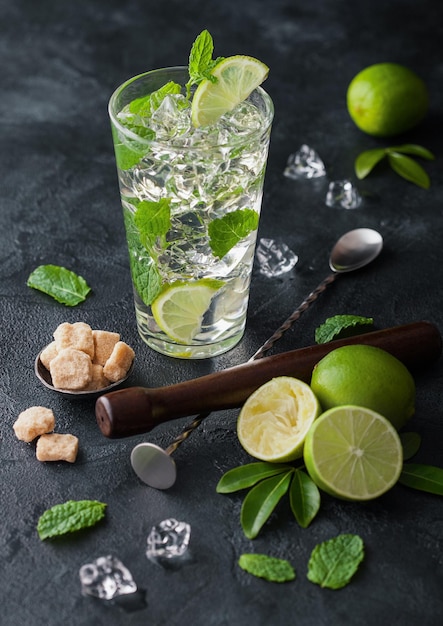 Glas Mojito-cocktail met ijsblokjes, munt en limoen op een zwart bord met lepel en muddler en verse limoenen en rietsuiker Beste cocktail voor de zomer