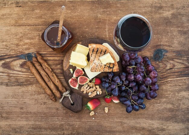 Glas rode wijn, kaasplank, druiven, vijgen, aardbeien, honing en brood sticks op rustieke houten tafel