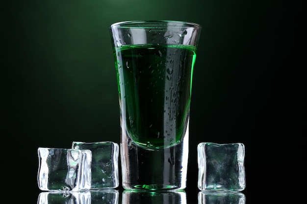 Glass of absinthe and ice on green background