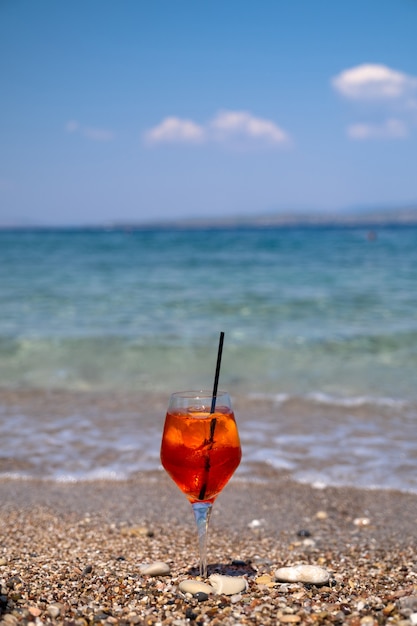 Glass of aperol spritz cocktail stands on sand near the sea