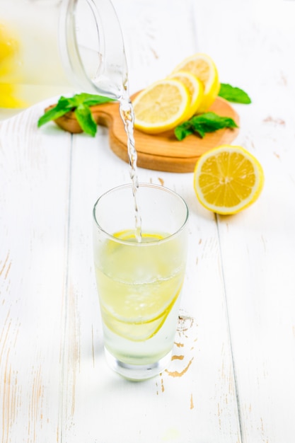 In a glass beaker, a cold lemonade is poured jug on white wooden background surrounded by lemons.