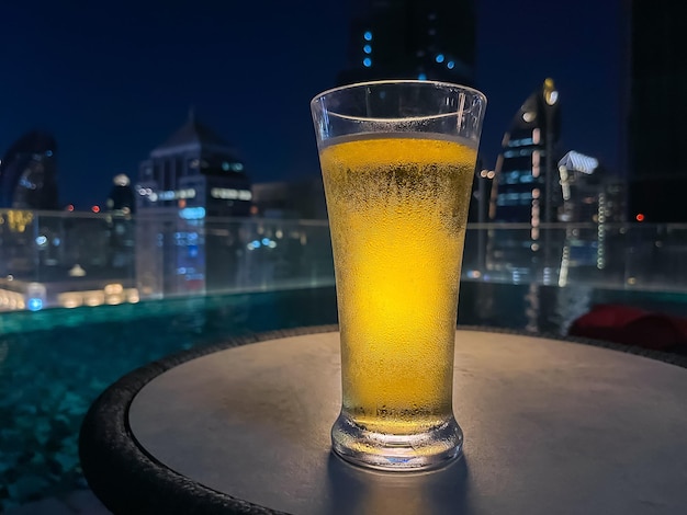 A glass of beer puts on table at rooftop bar with colorful city\
background