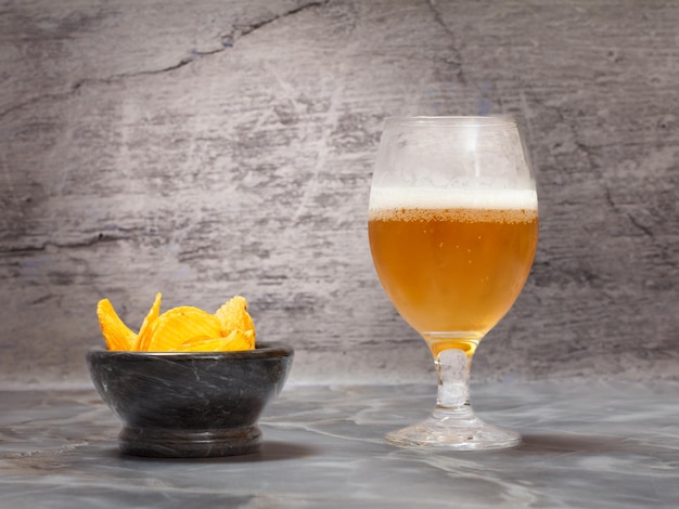 Glass of beer and a stone bowl with potato chips on the gray background.