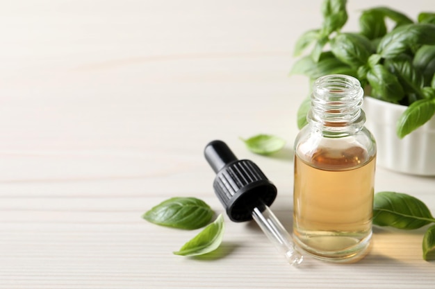 Glass bottle of basil essential oil and leaves on white wooden table Space for text