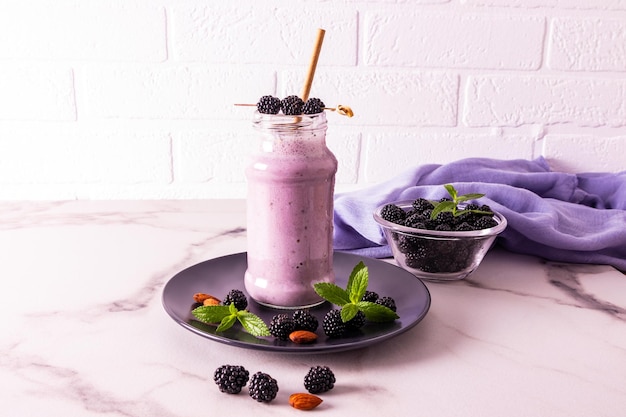 A glass bottle of blackberry smoothie or cocktail stands on a dark ceramic plate on a marble table against a brick wall healthy eating diet