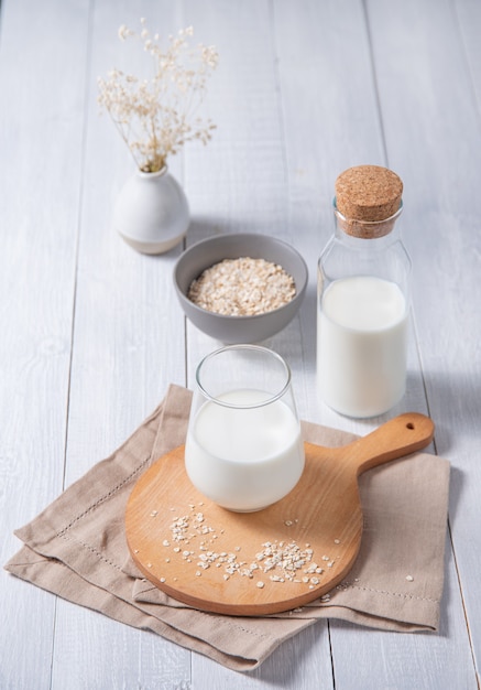 Foto un bicchiere e una bottiglia di latte di farina d'avena fresca e alcuni fiocchi di farina d'avena sparsi su un fondo di legno bianco