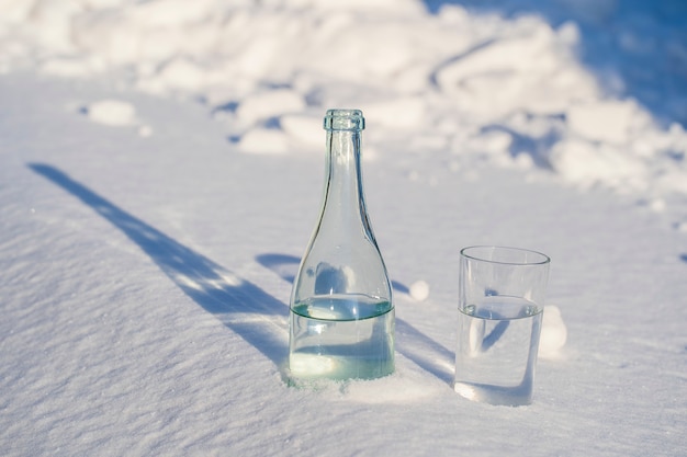 Стеклянная бутылка и стакан воды на белом снегу зимой