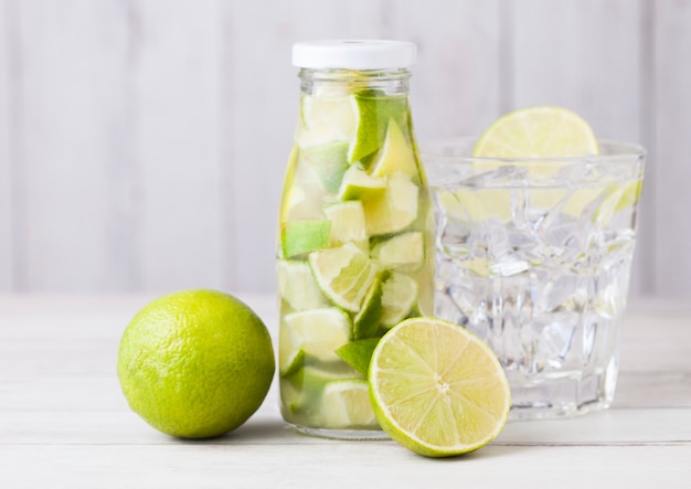 Glass bottle of lime slices still fruit water and raw limes on white wooden with glass and ice cubes