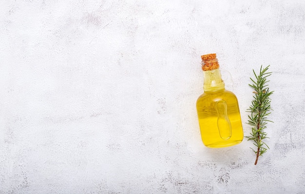 Glass bottle of olive oil and rosemary branch set up on white concrete background.