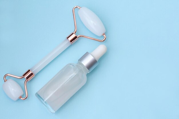 A glass bottle with a nonbranded serum and a face massage roller on a blue background top view