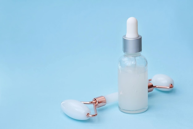 A glass bottle with a nonbranded serum and a face massage roller on a blue background