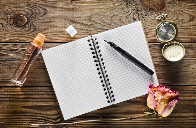 A glass bottle with perfume, notebook and a pen, dry rose, vintage pocket watch on a rustic wooden table. Top view.