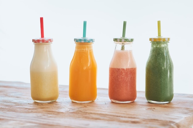 Glass bottles with colorful straws filled with assorted fruit smoothies served on wooden table in row in light room