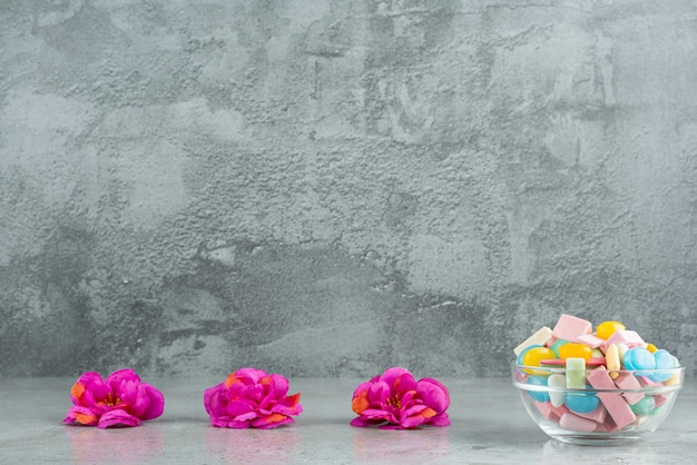 Glass bowl of colorful chewing gums with flowers on stone surface.