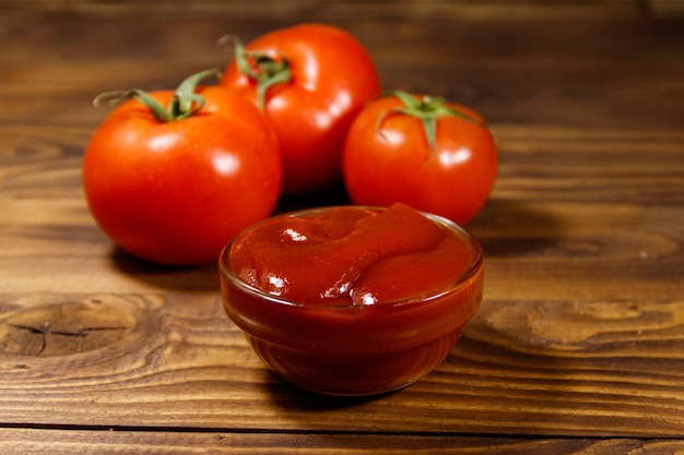 Glass bowl of ketchup or tomato sauce and fresh ripe tomatoes on wooden table