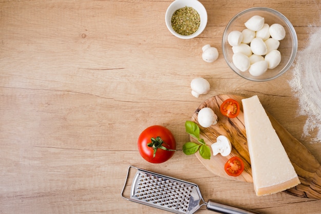 Glass bowl of mozzarella cheese; tomatoes; basil and cheese block with grater on desk